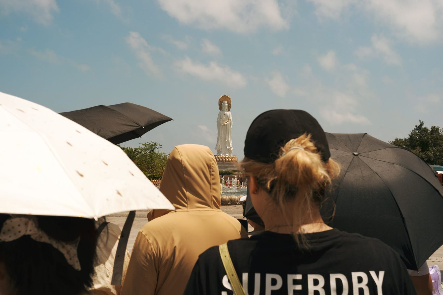 Tourist group with sun umbrellas and person wearing SUPERDRY shirt. Guanyin statue peeking through in the distance.
