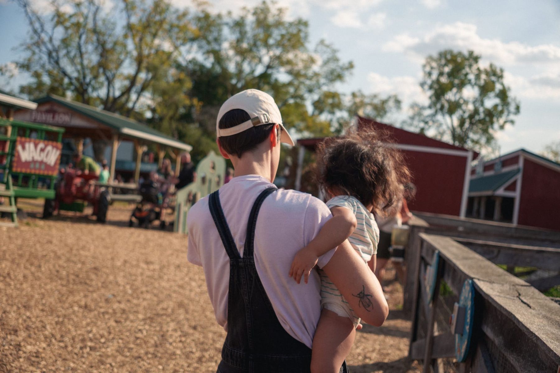 Back angle shot of Julia carrying Rufus, outdoors with small farm buildings and wood chip ground.