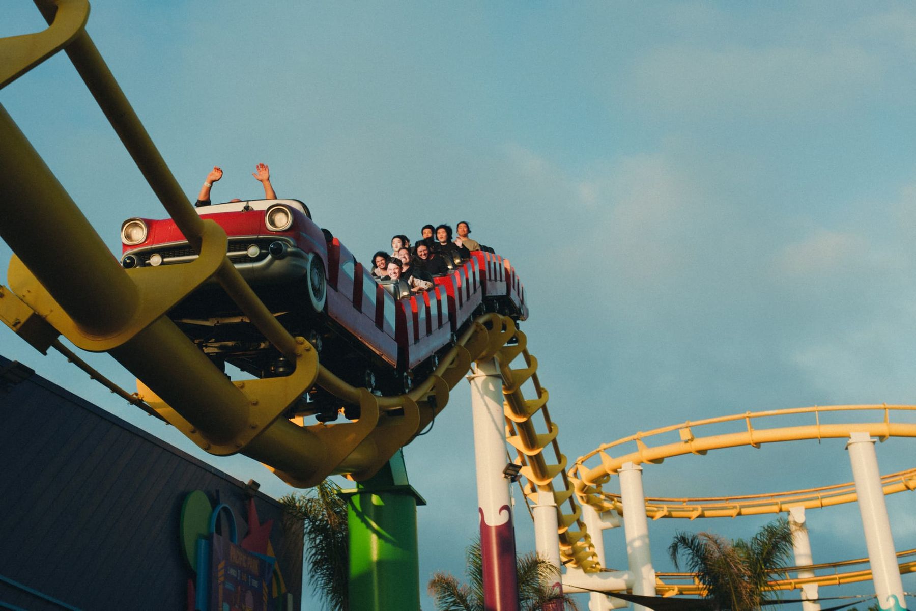 Ground-level view of roller coaster riders at top of track, palm trees and cloudy blue-gray sky.