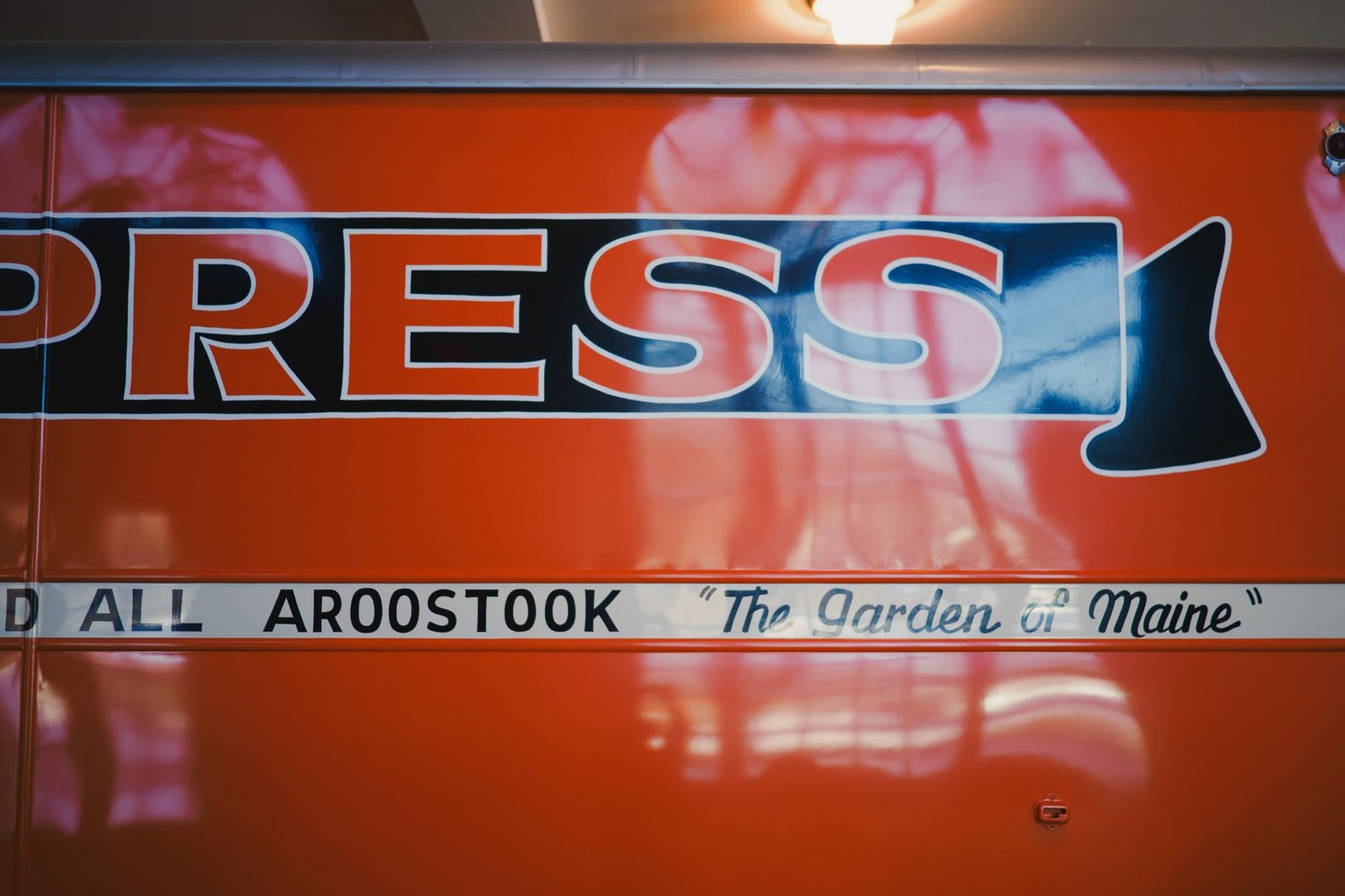 Window reflections off an orange EXPRESS bus, visible words “ALL AROOSTOOK, ‘The Garden of Maine’”