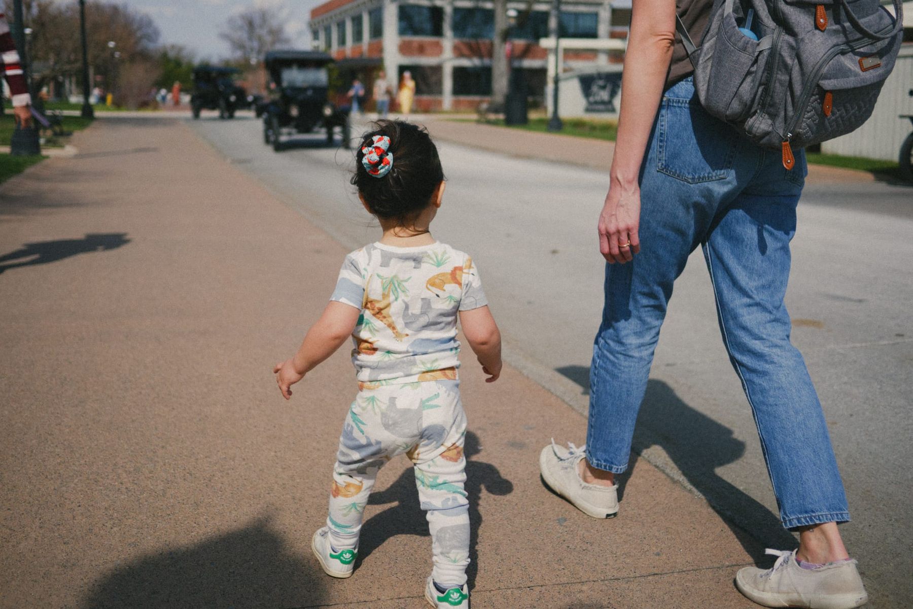 Toddler in a dinosaur-print outfit toddles down a sunny brick street, seen from behind, with an adult in jeans walking alongside. Vintage cars in the background.