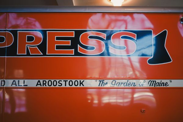Window reflections off an orange EXPRESS bus, visible words “ALL AROOSTOOK, ‘The Garden of Maine’”