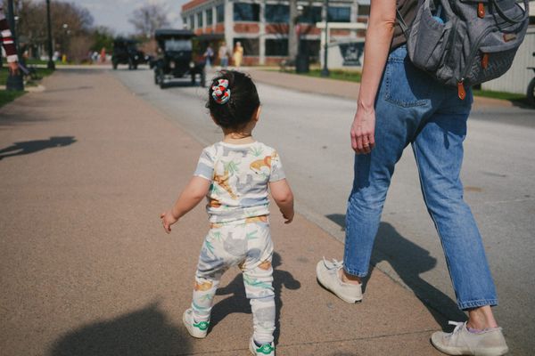Toddler in a dinosaur-print outfit toddles down a sunny brick street, seen from behind, with an adult in jeans walking alongside. Vintage cars in the background.