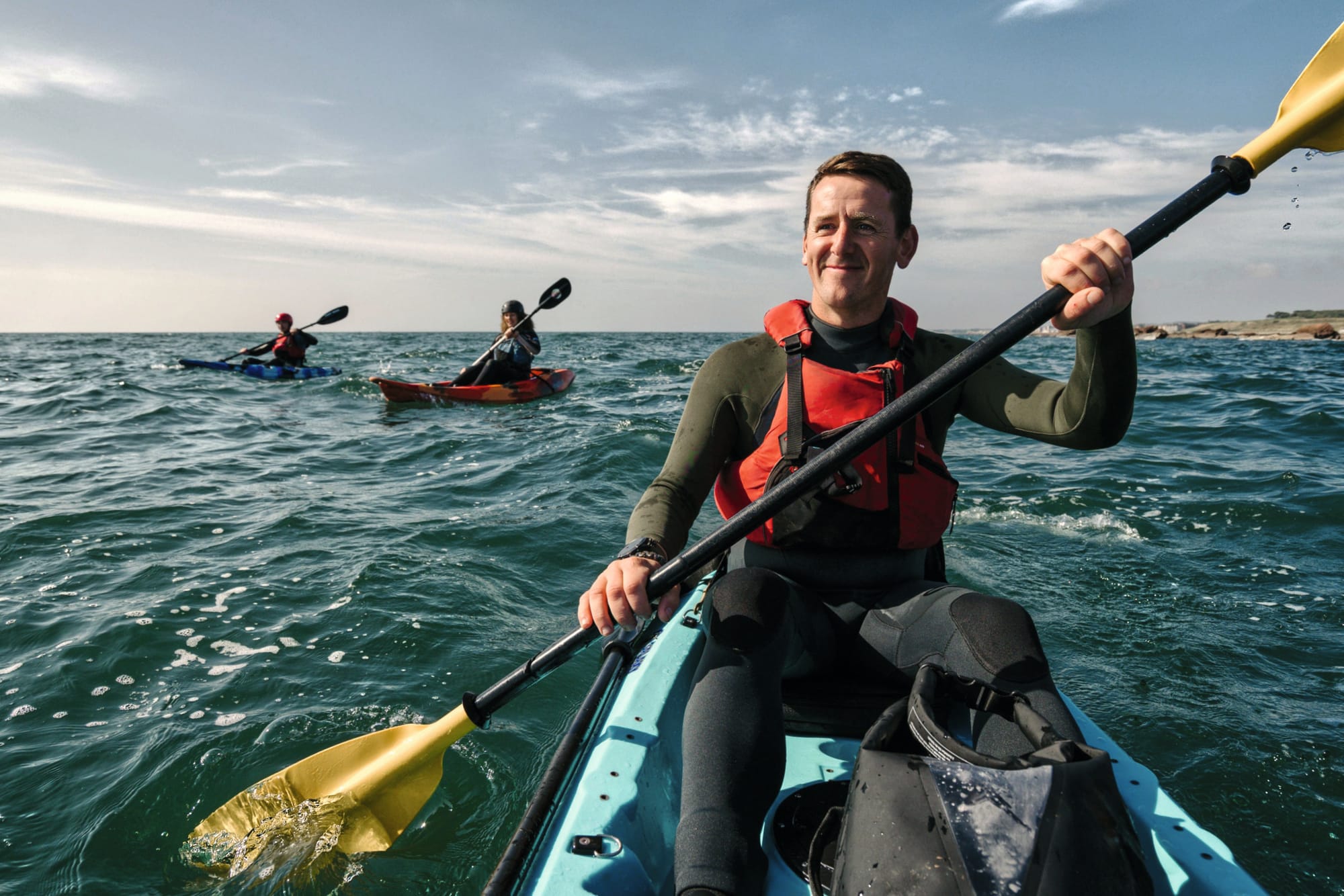 Cam in a kayak out on the sea holding an oar. There are two more kayaks in the background.