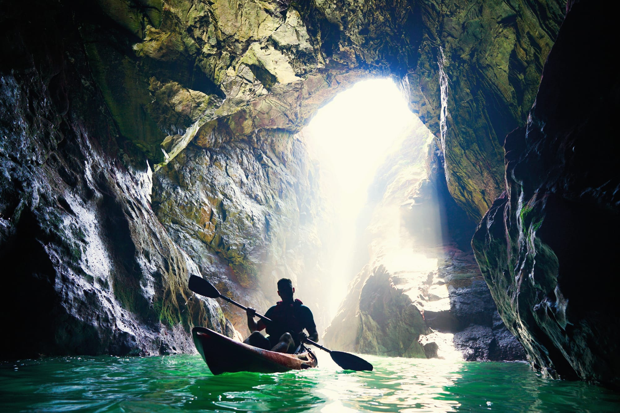 The water in the cave is green. A man is kayaking and there is light coming through from a hole in the top of the cave.