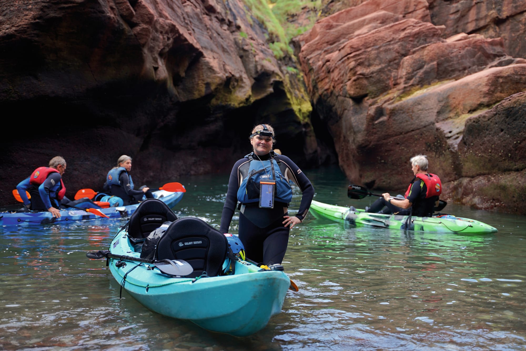 Jacki standing next to her kayak in shallow water. Caves in the cliffs are behind her.