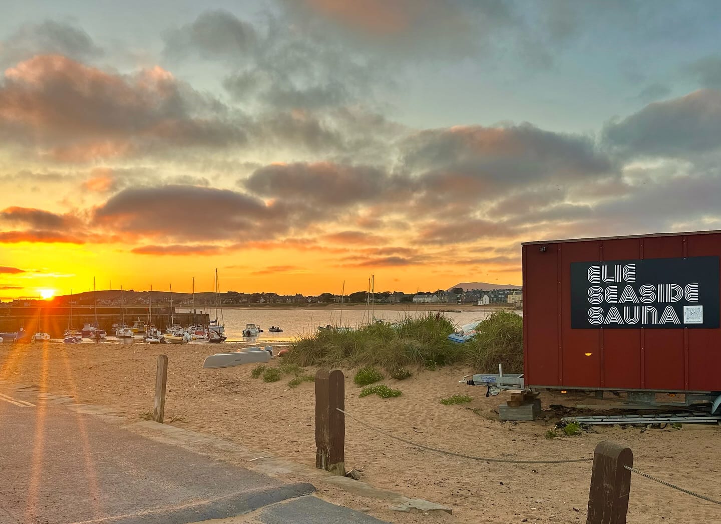 Shore, with a sign reading "Elie Seaside Sauna", is located on the beach by the harbour. The sun is setting and the water is in the background.