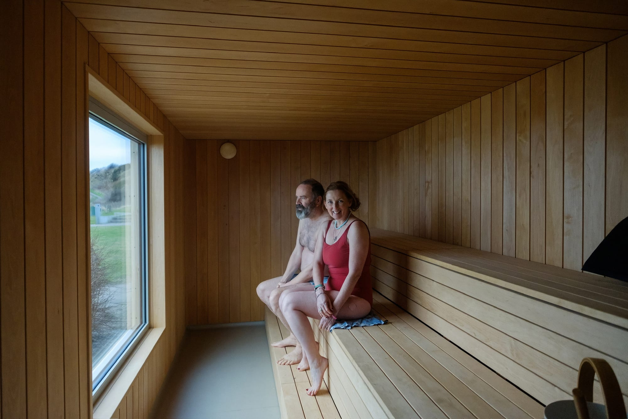 St Andrews Seaside Sauna owners Greg and Julie sit on the wooden bench in the sauna. There is a window looking out to the right of the image.