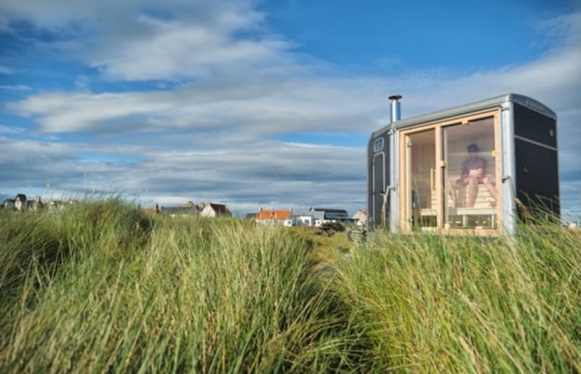 Dune sauna sits within long grass. People are visible sitting in the sauna, and the town of Elie can just be seen in the background.