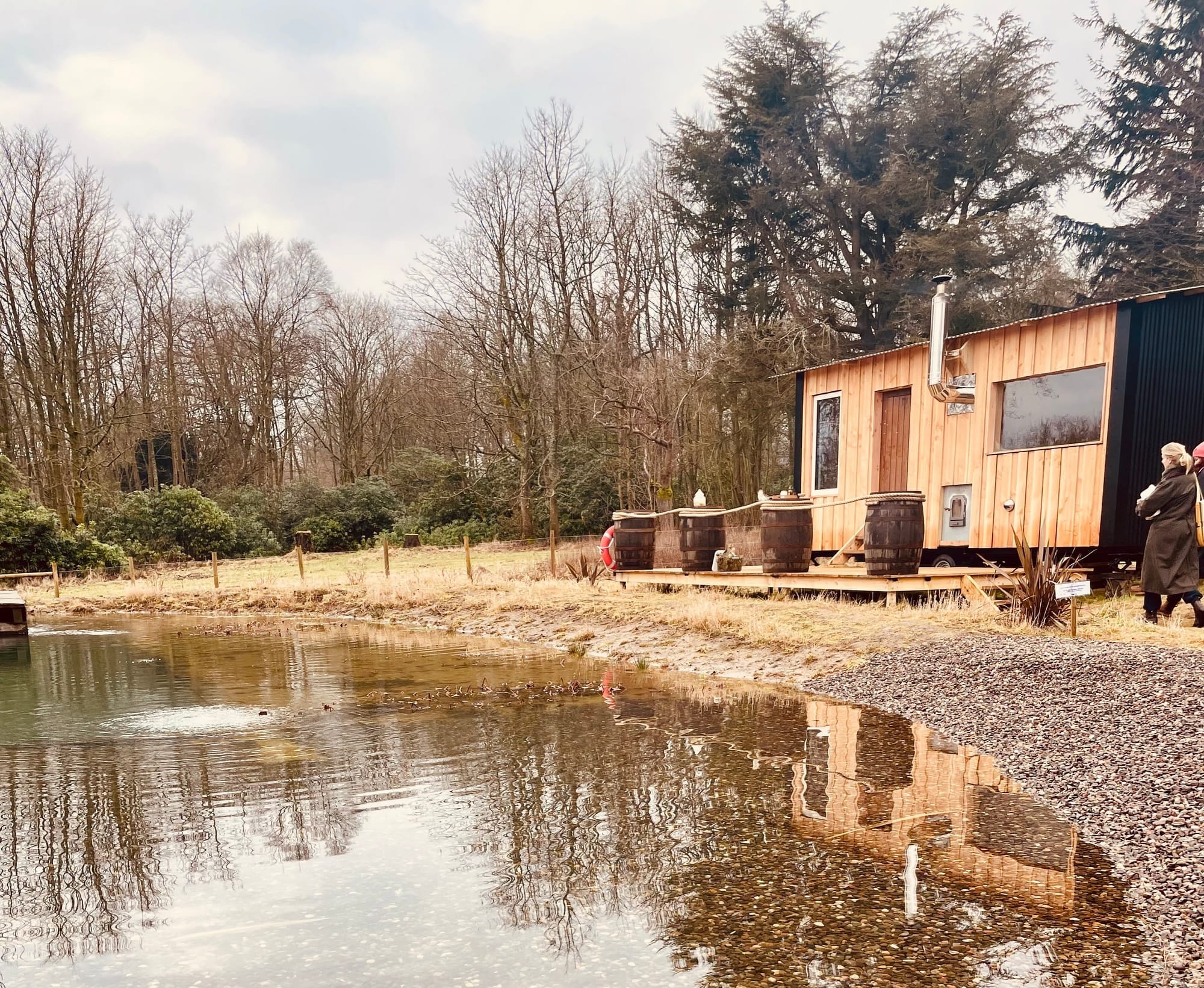 A wooden cabin sits on grass surrounding a natural pool. Trees surround the cabin in the background.