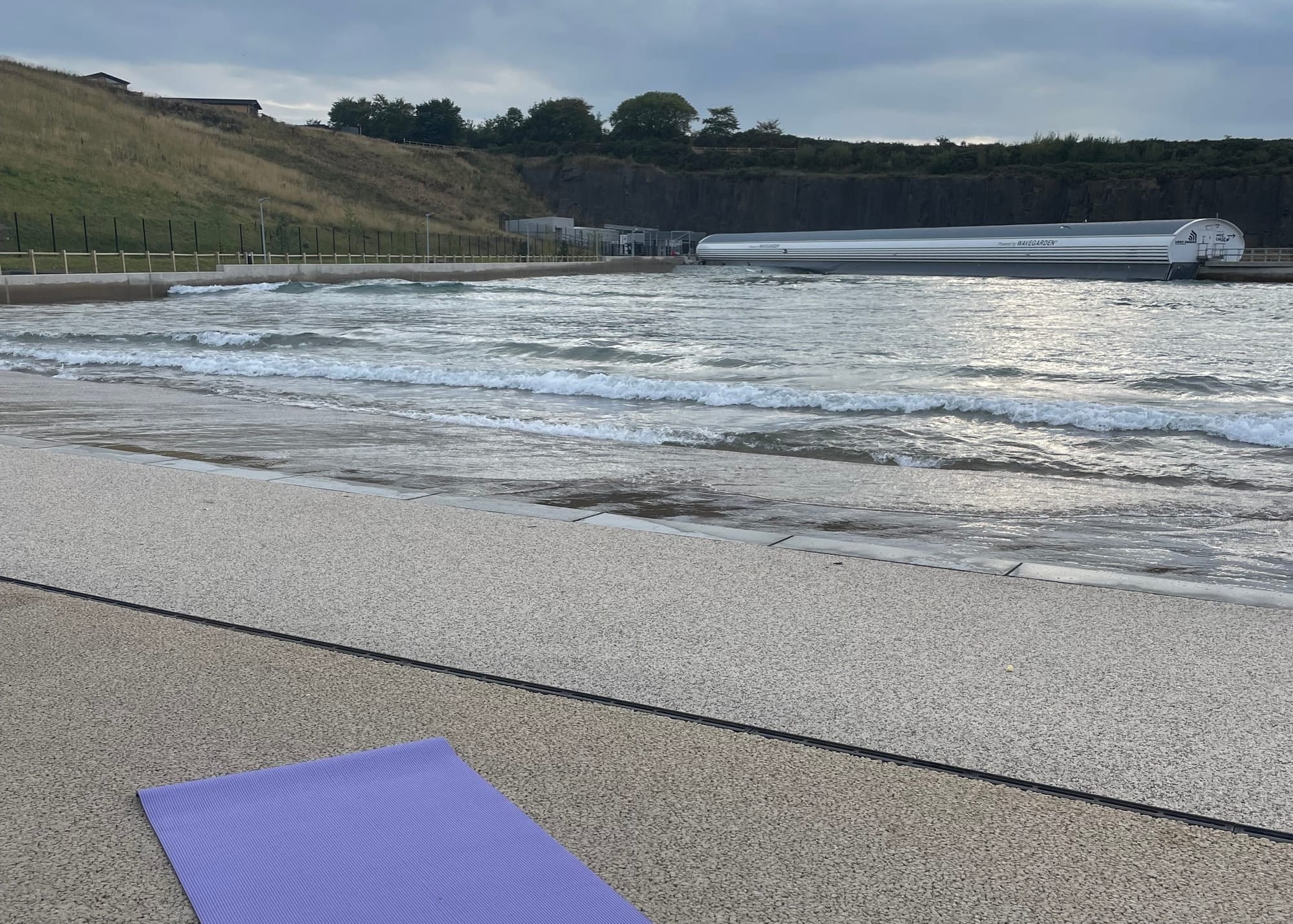 A purple yoga mat lies in the foreground, with the water of the concrete surf lagoon behind it. 
