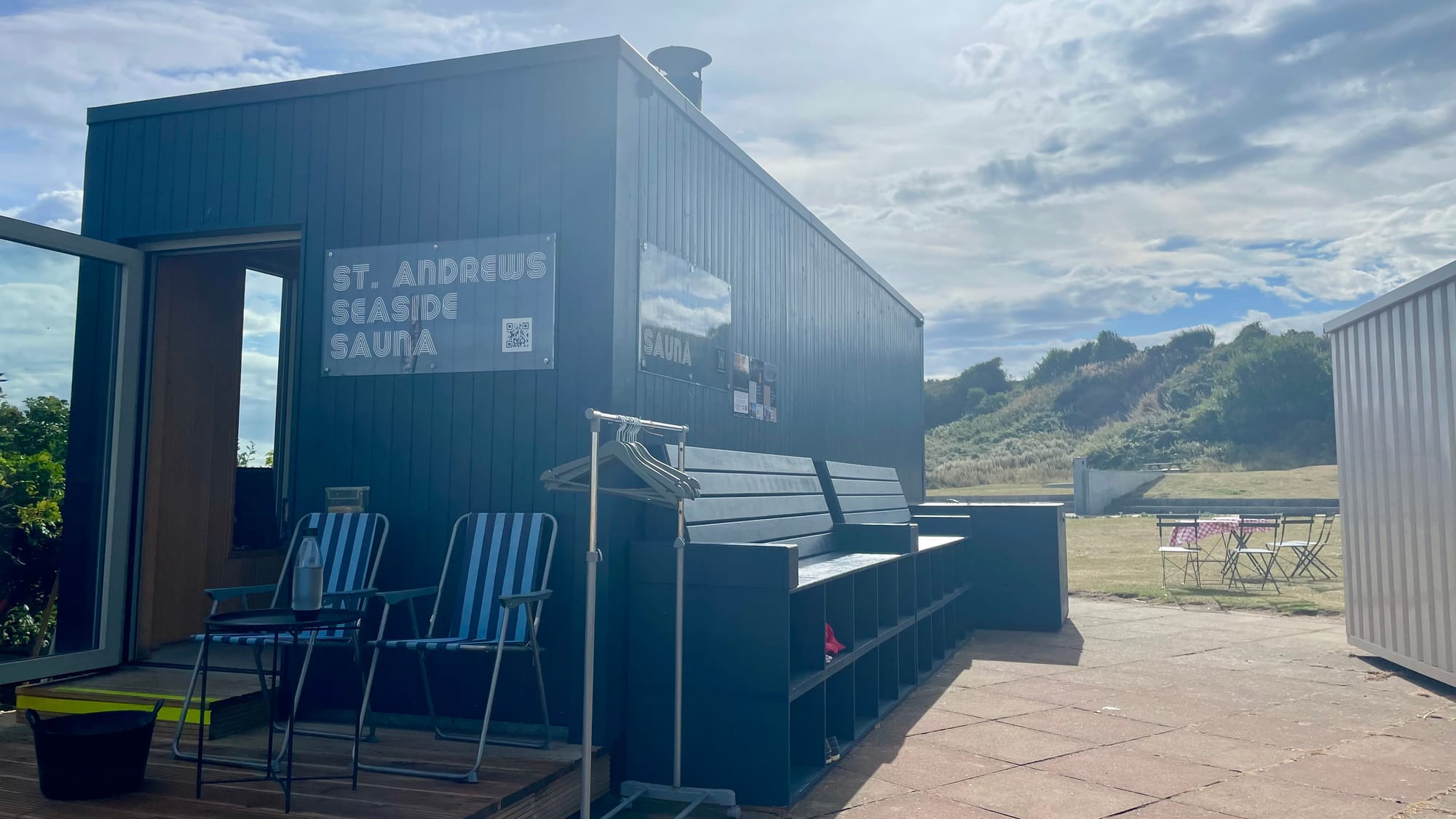 St Andrews Seaside Sauna from the outside. The building is black wood panelling and it is situated on paving stones with a green stretch of grass in the background. 