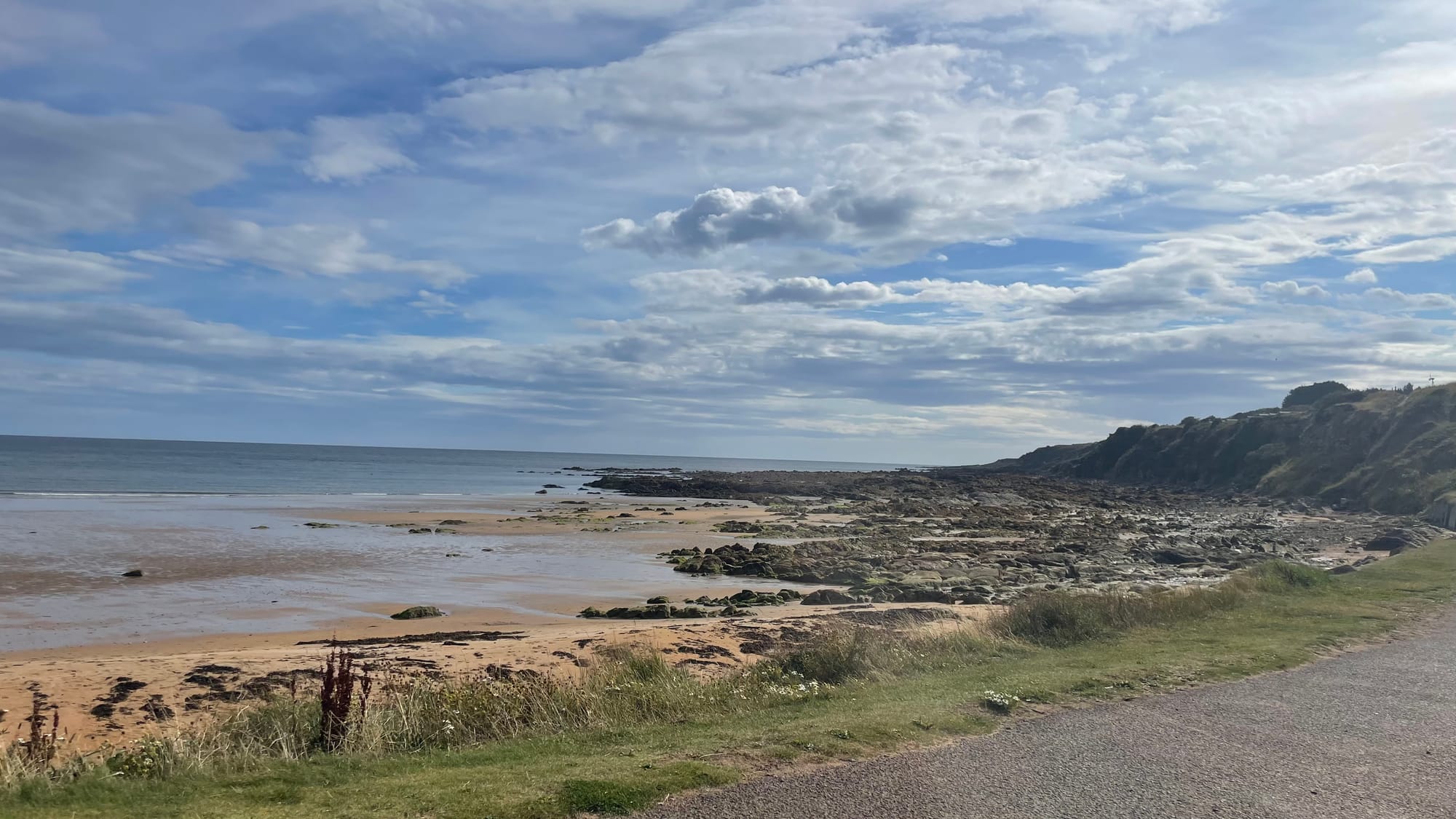 View across East Sands beach. A grassy verge leads onto the sand. The far end of the beach is very rocky and the tide is in.