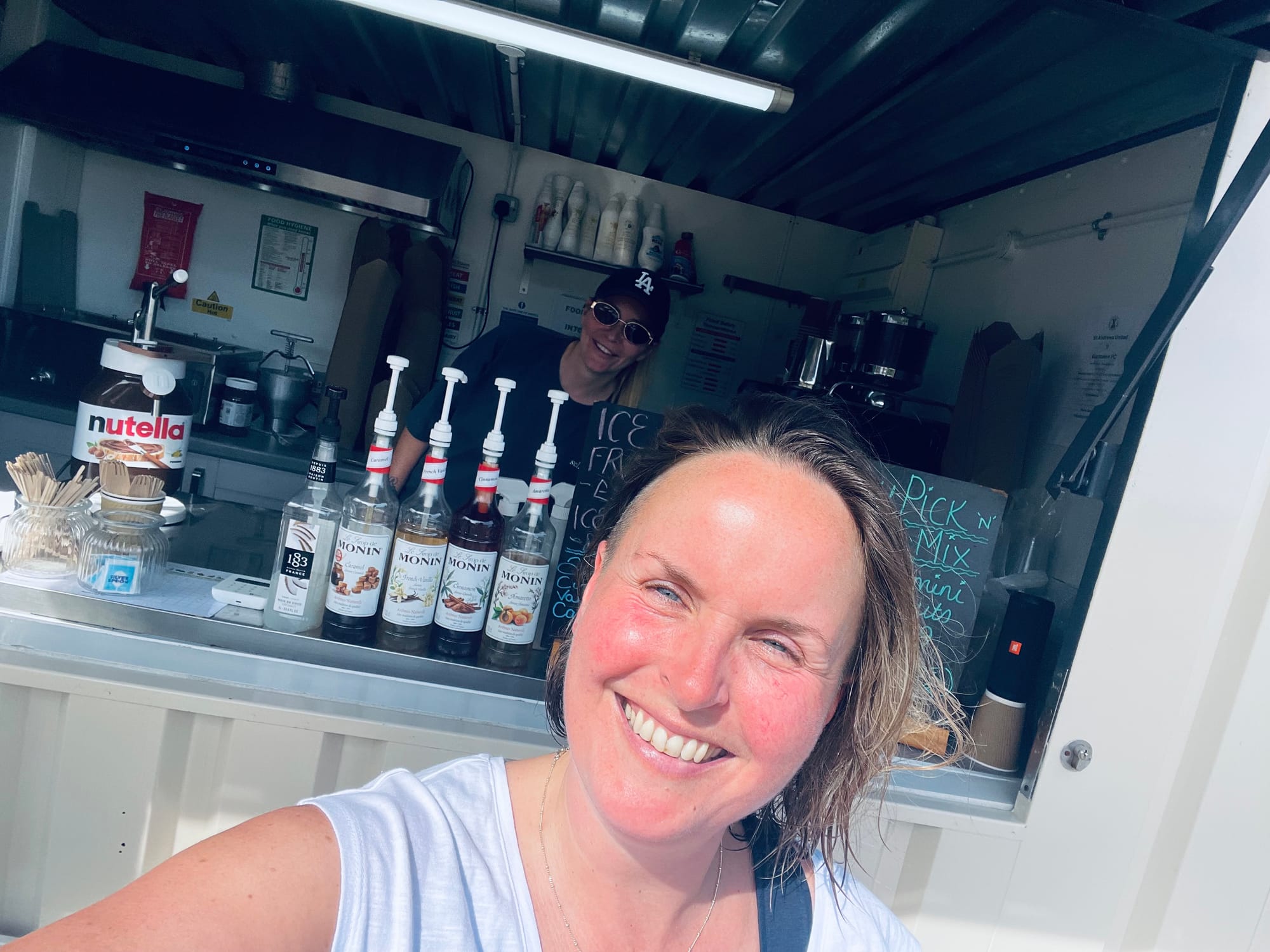 Rachel smiles with the worker in Caffe Bombolino. Coffee syrups, menus and disposable cutlery line the shelf.