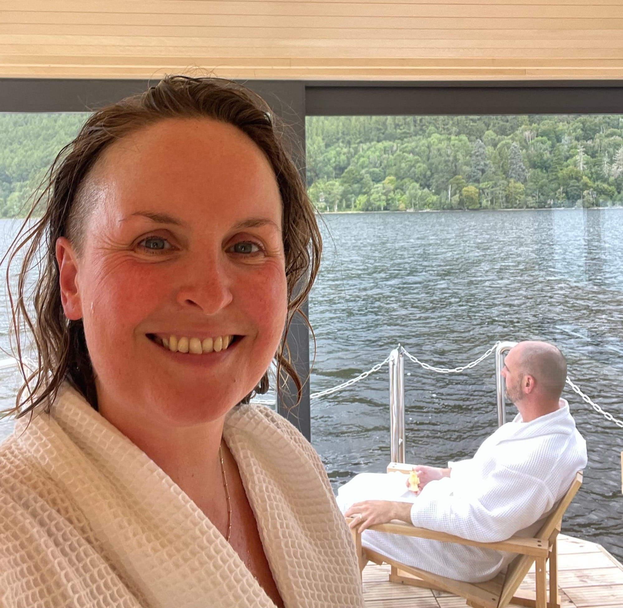 Rachel smiles for a selfie with Kieran sitting in a wooden chair in the background. They are overlooking the water from the sauna cruise.