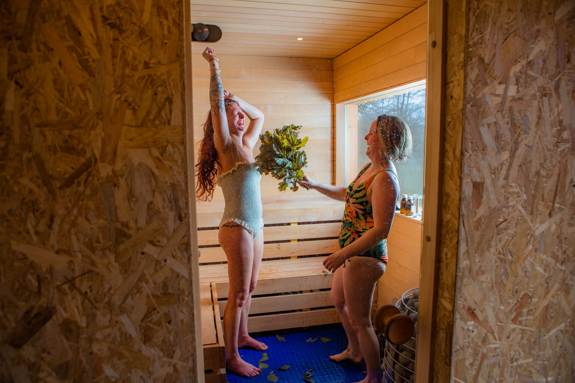 Inside the sauna, Rachel is holding an oak whisk up to Katrino.