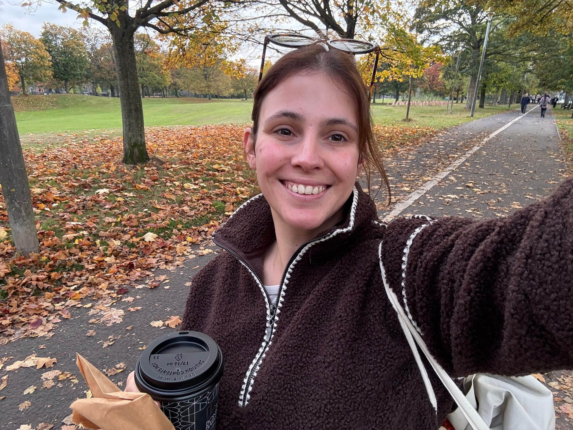 Daniella smiles for a selfie on a path through a park. There are fallen leaves covering the grass behind her.
