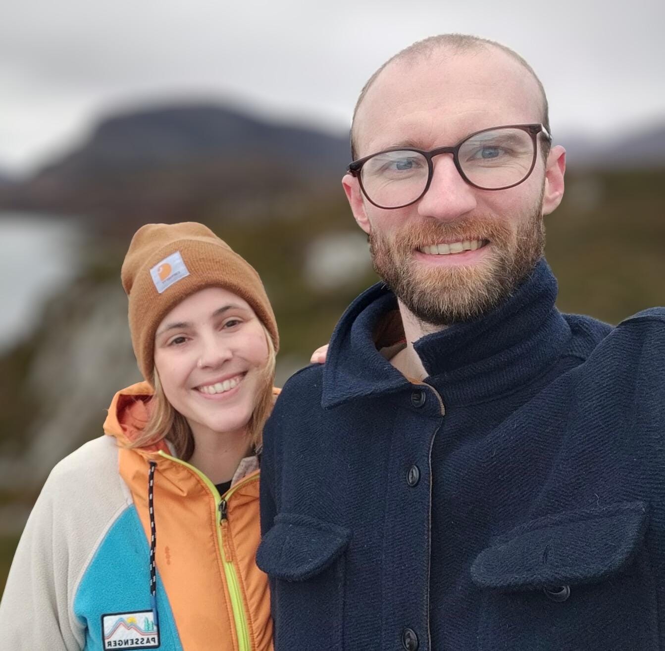 A young woman in a warm hat and coat stands smiling with her husband who wears a thick navy blue coat.