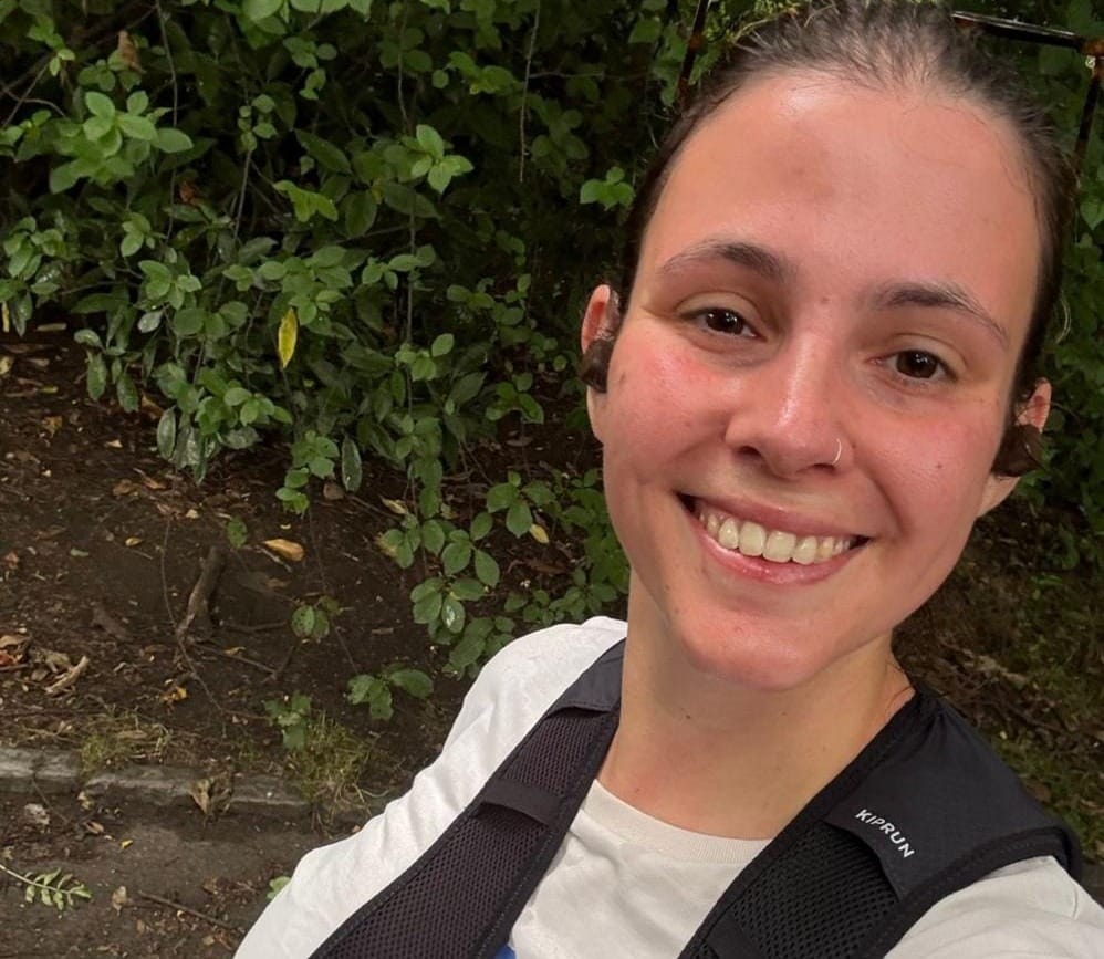 Daniella smiles for a selfie on one of her runs. There is a green bush behind her.