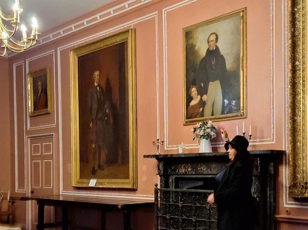 Lynne standing next to the fireplace in the Pink Room. There are large portraits on the wall. 