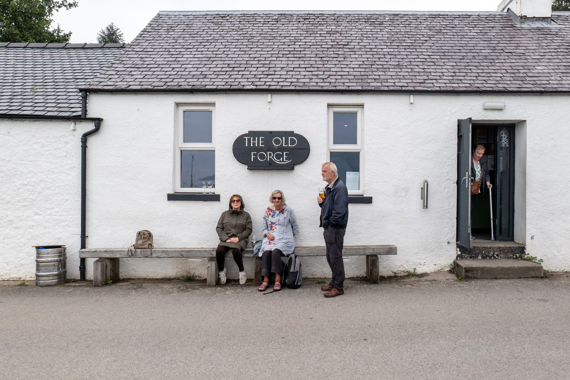 People sit on a bench outside The Old Forge pub, which is painted white with a grey tiled roof.