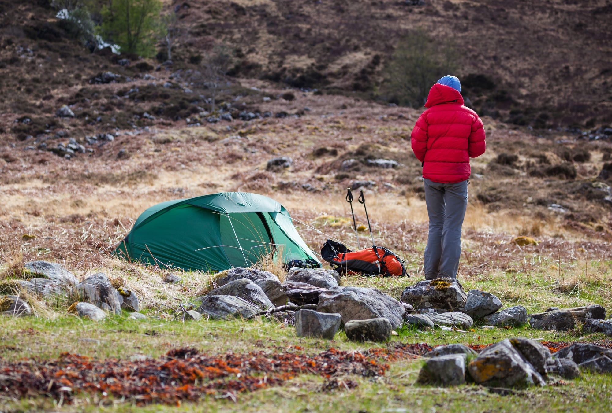 A small green tent on grass. A hiker stands next to it in a red puffer coat, with a rucksack and hiking poles laid next to the tent.
