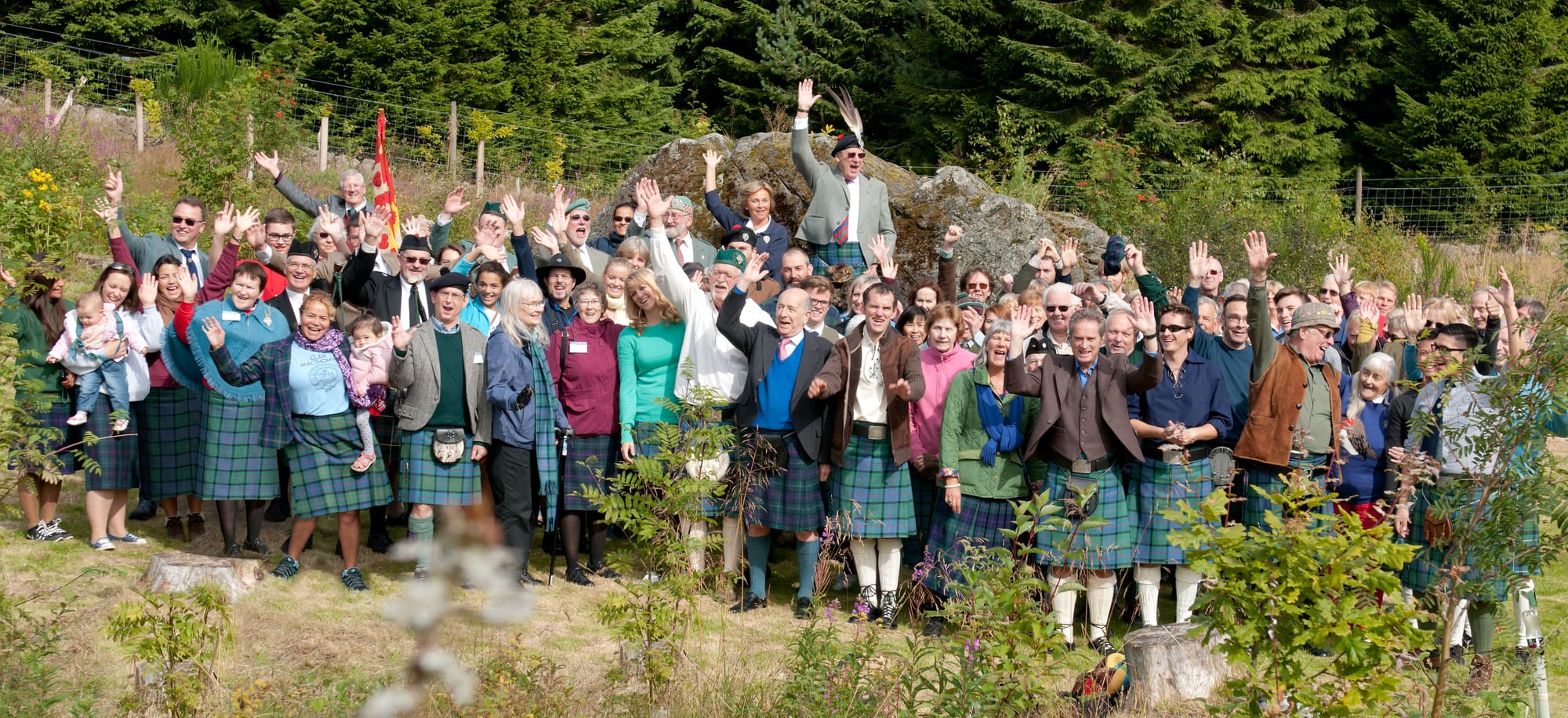 A large group of people, many wearing green kilts, stand cheering in front of a large rock by a forest.