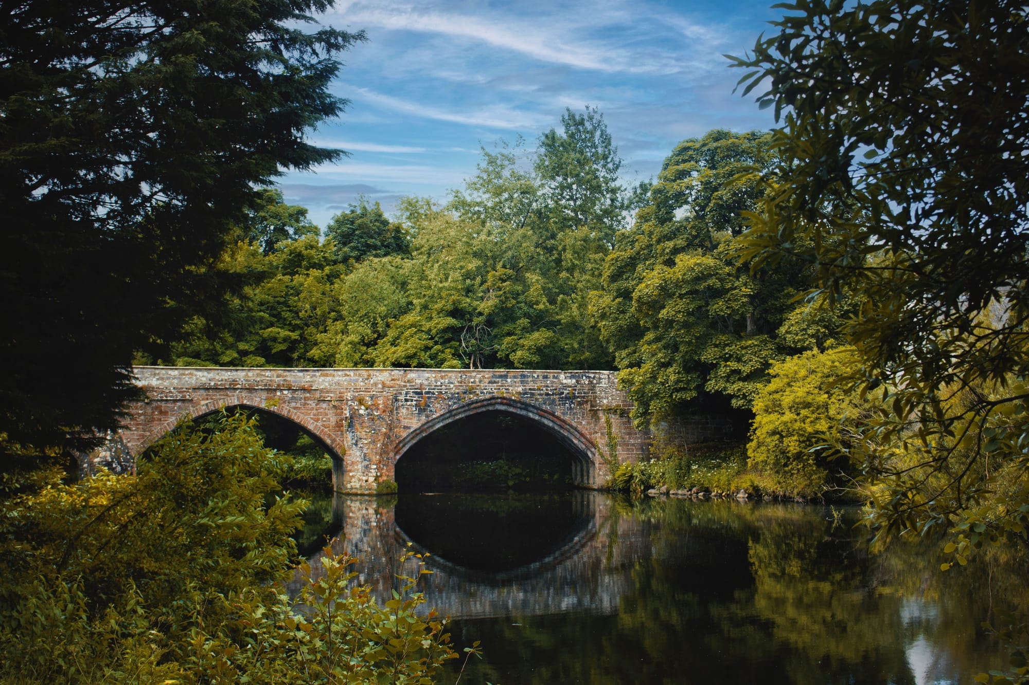 An old stone bridge over a still river surrounded by green trees lining the banks.