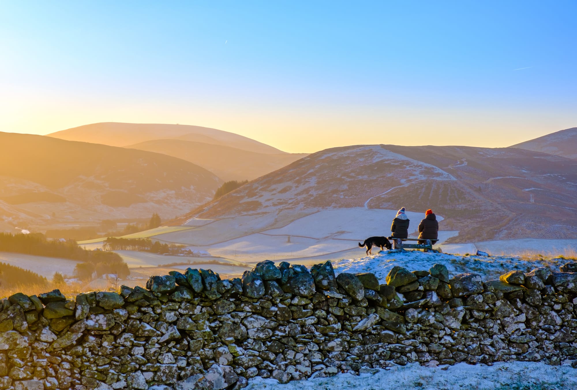 Two people sitting on a bench with a dog next to them, looking out over snowy hills. There is a low wall in the foreground.