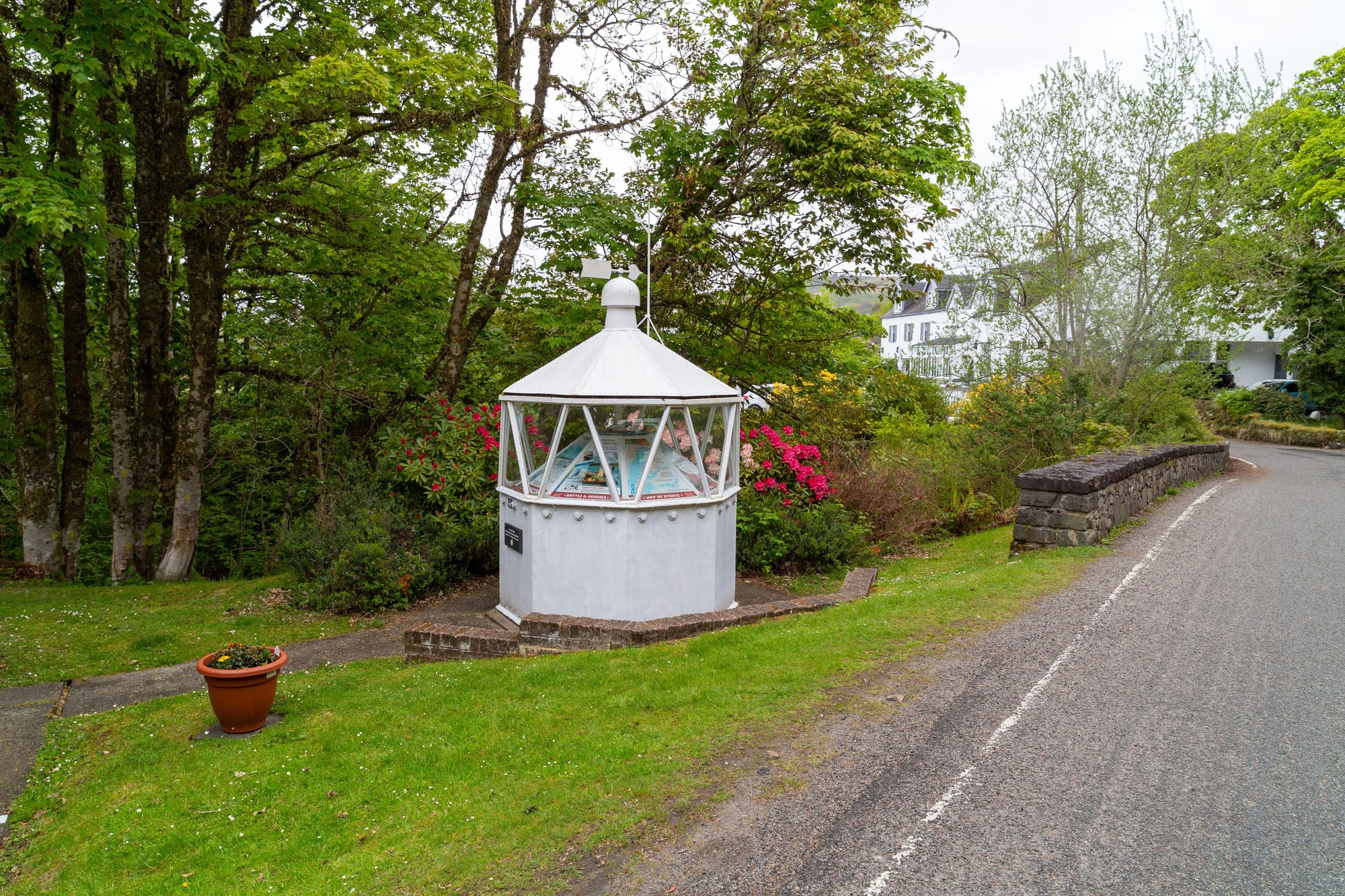 A white lighthouse lantern sits by a path and a well-maintained patch of grass at the side of a road..