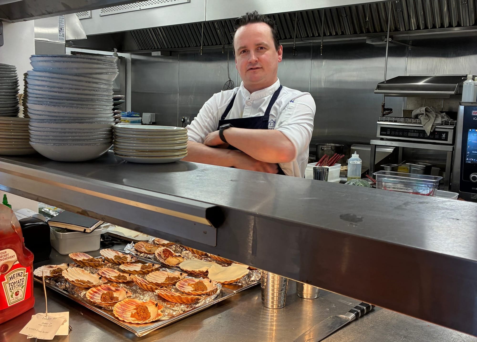 A male chef stands behind the pass with his arms folded.