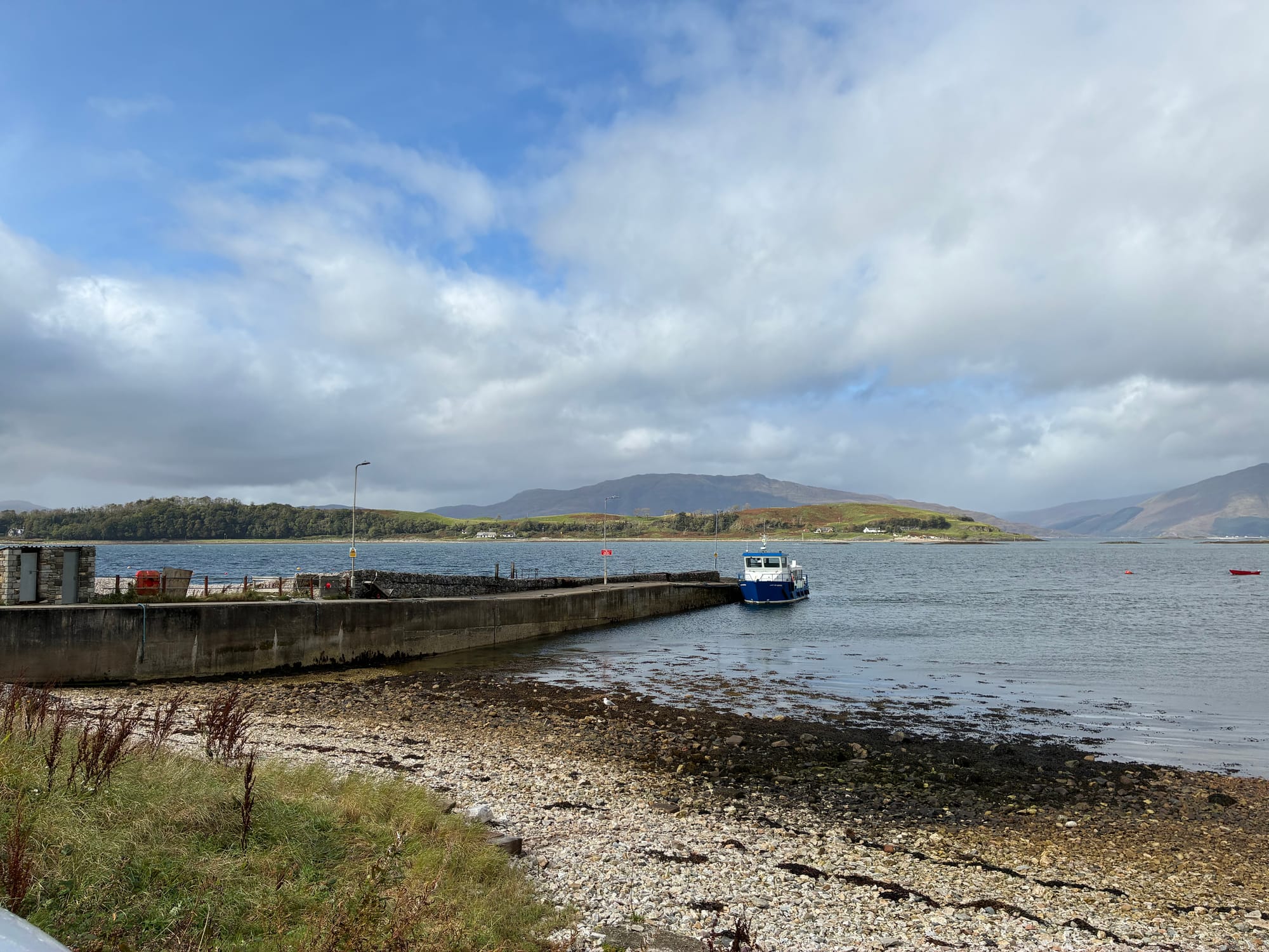A concrete jetty stretches out into a sea loch with a small blue and white ferry waiting at the end of it to go to the little island in the distance.