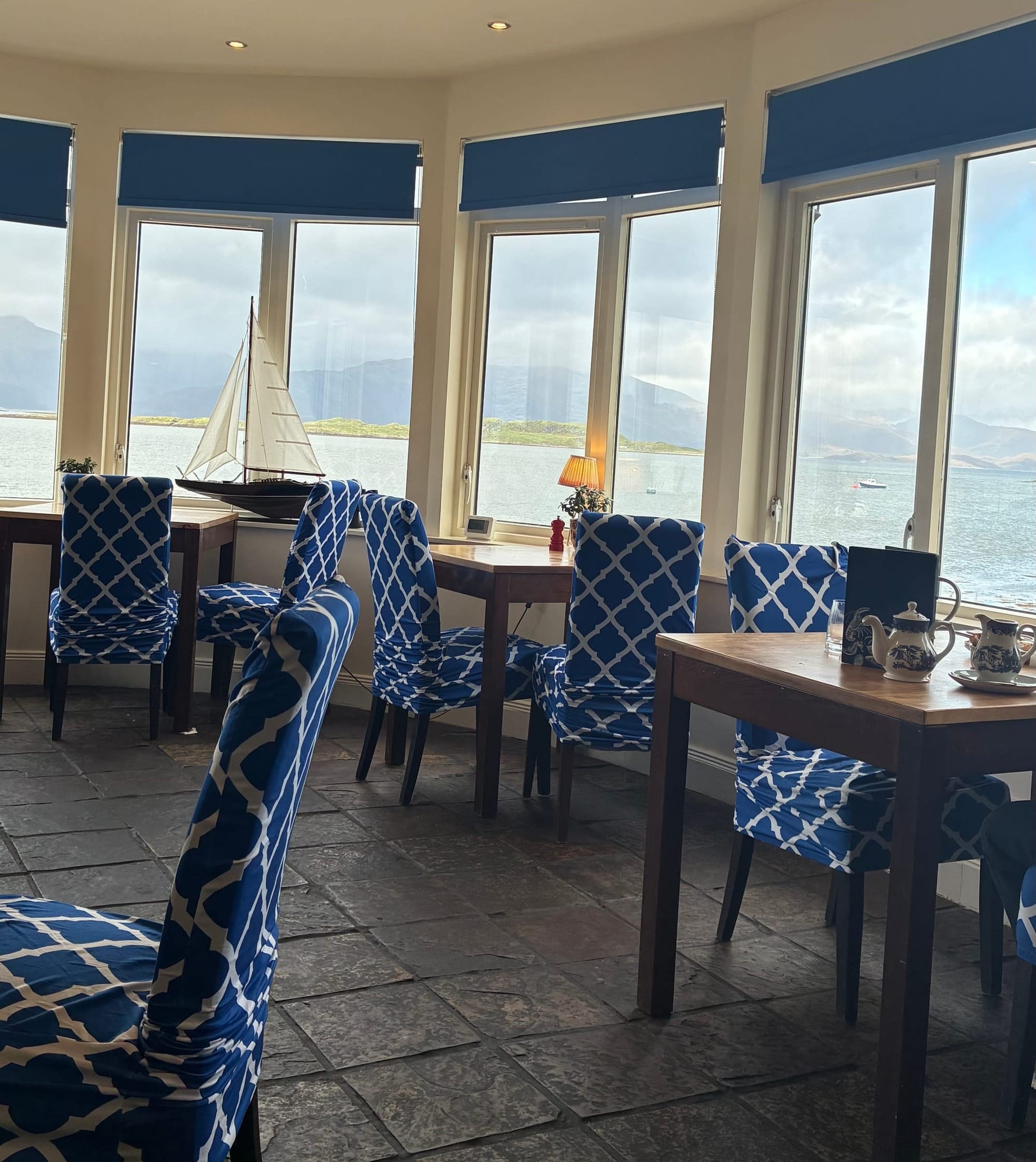 A restaurant with blue and white high-backed chairs and many windows looking out to the loch and mountains in the distance.