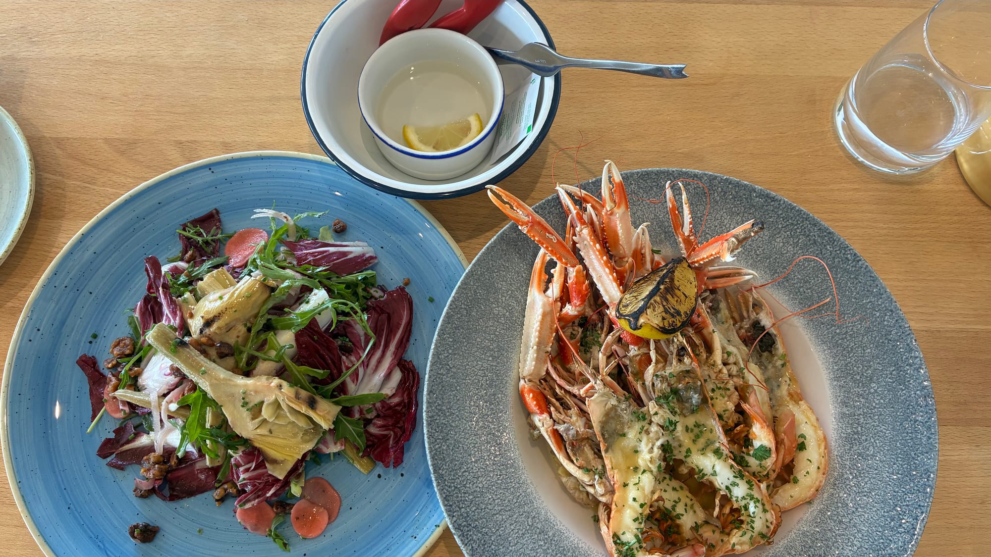 Two blue round plates of grilled langoustines and an artichoke heart salad on a wooden table.