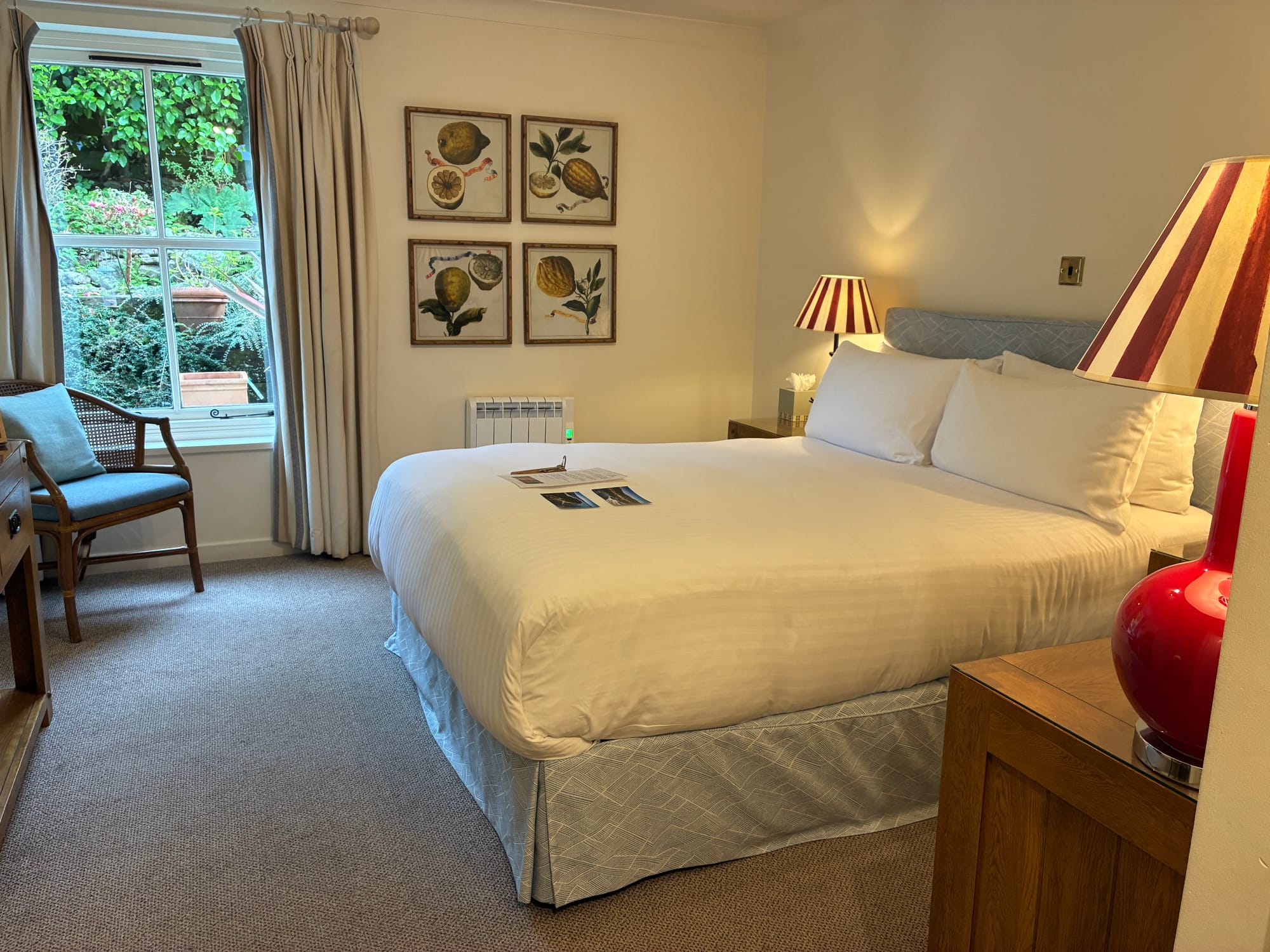 A cost hotel room with comfy white bed, red and white striped bedside lamps and a window looking out to the garden.