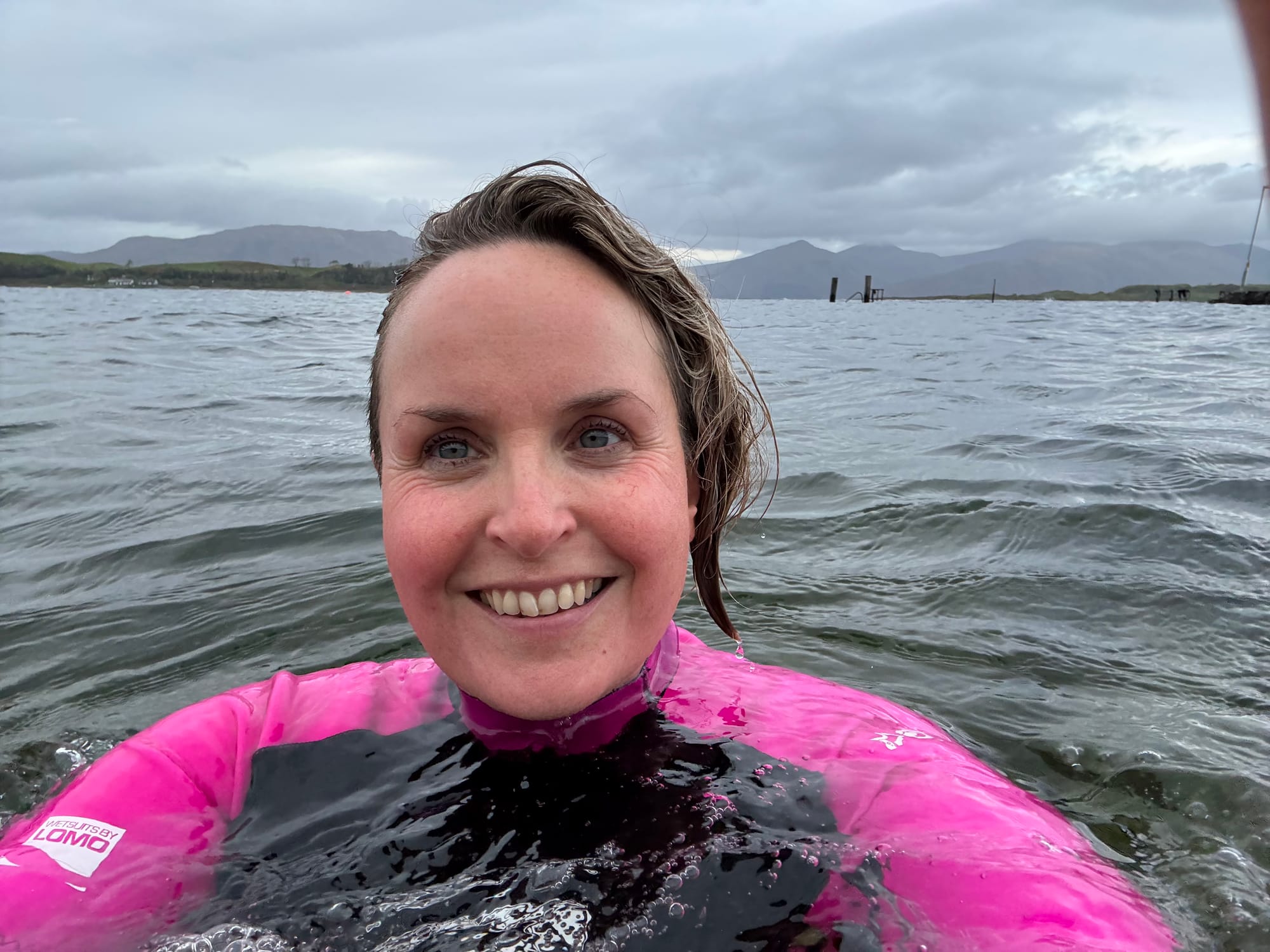 A woman in a pink and black wetsuit smiles in a cold loch while smiling at the camera.