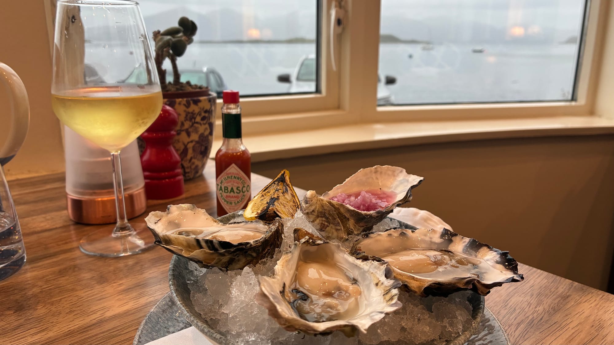 A round grey dish of crushed ice with four oysters on top next to a chilled glass of wine and a window with loch views.