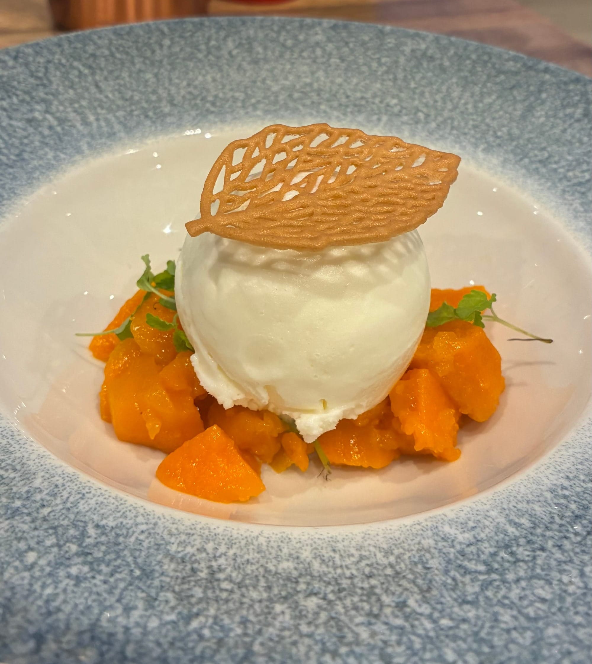A dessrt of chunks of opaque orange jelly topped with vanilla ice cream and a delicate leaf shaped wafer on top.