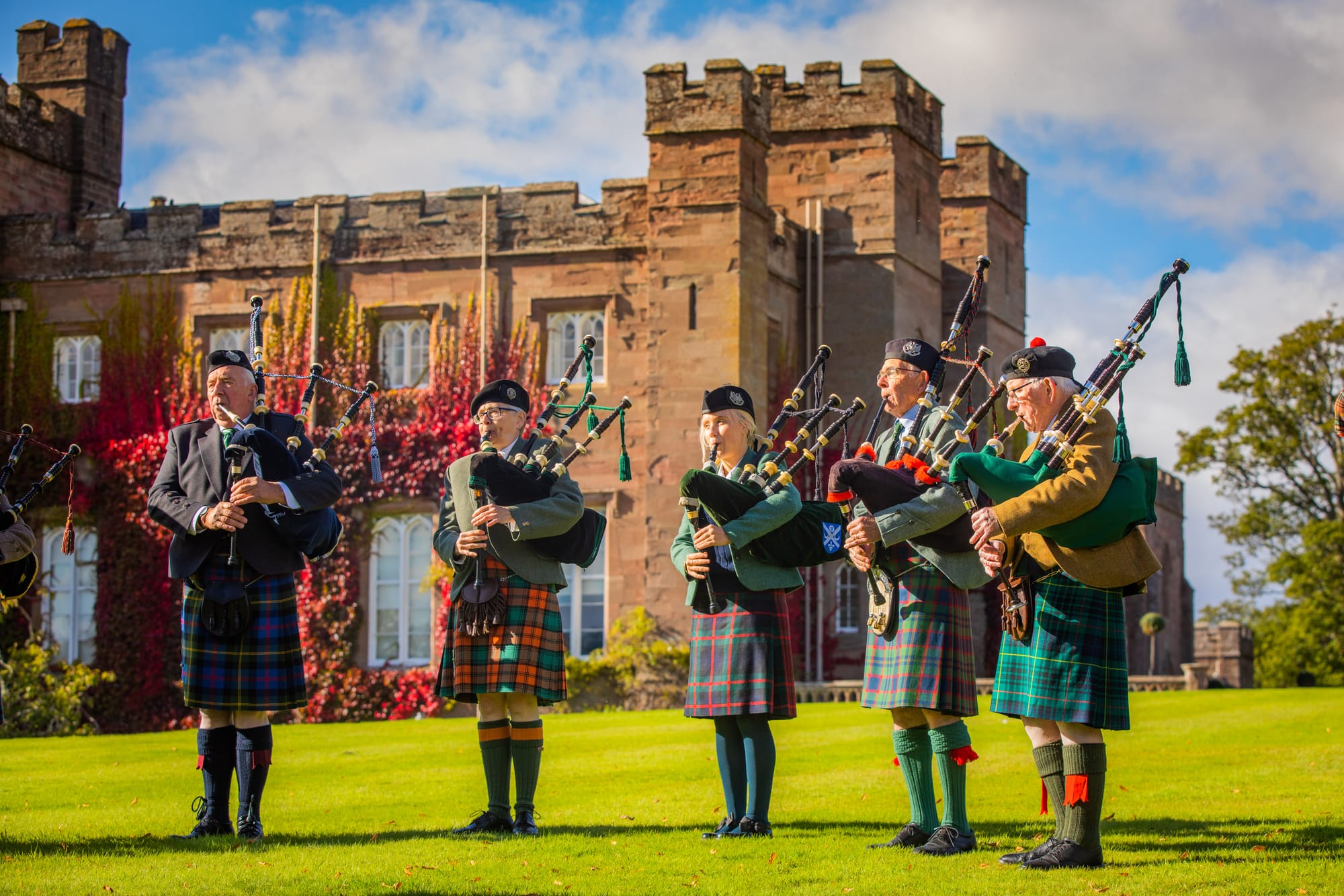 Four men and one woman playing bagpipes outside Scone Palace which is draped in reddish autumnal leaves.