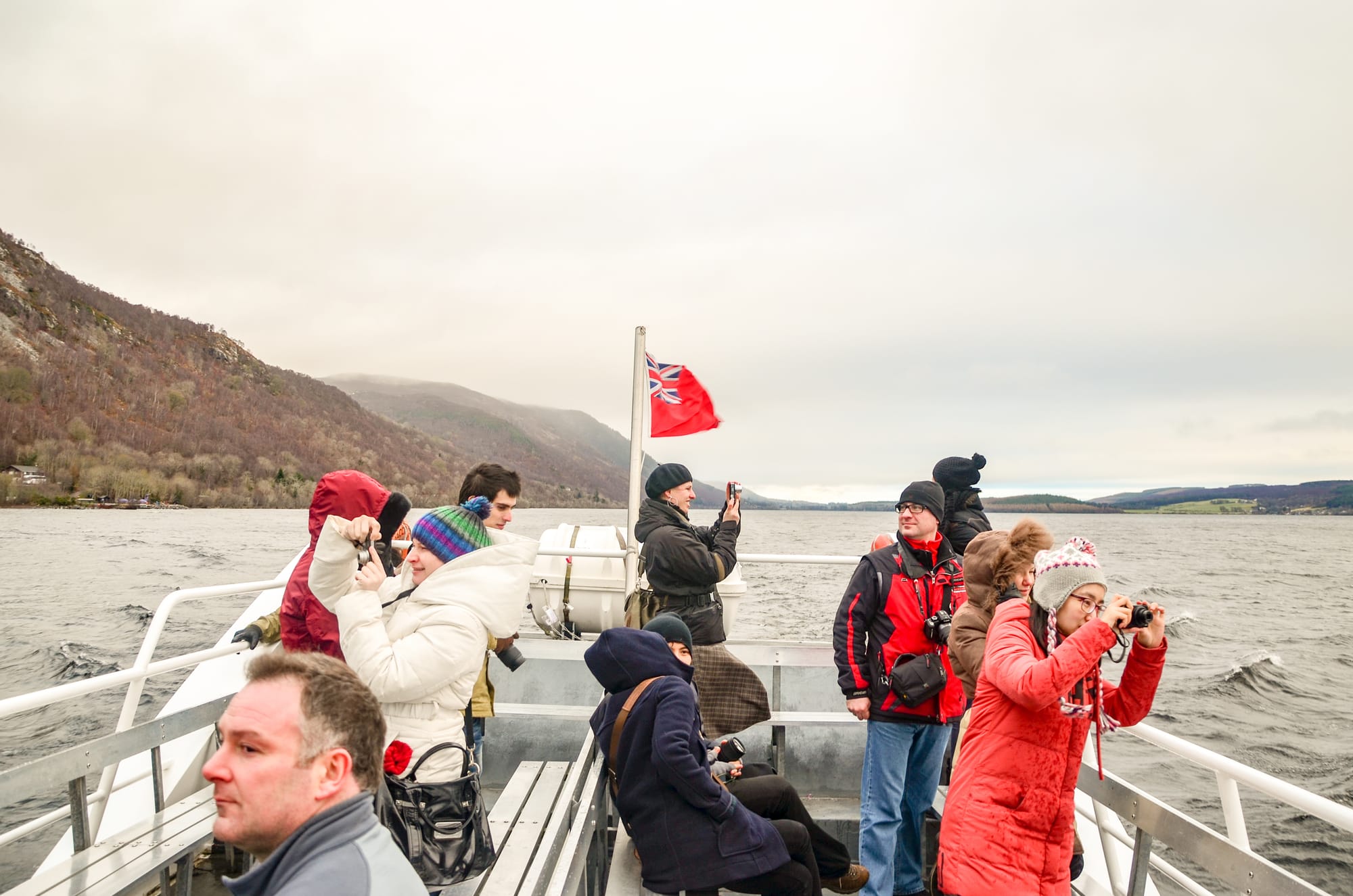Tourists on a boat tour of Loch Ness. They are well wrapped up in big coats and hats, and they are taking photos of the choppy, grey water.