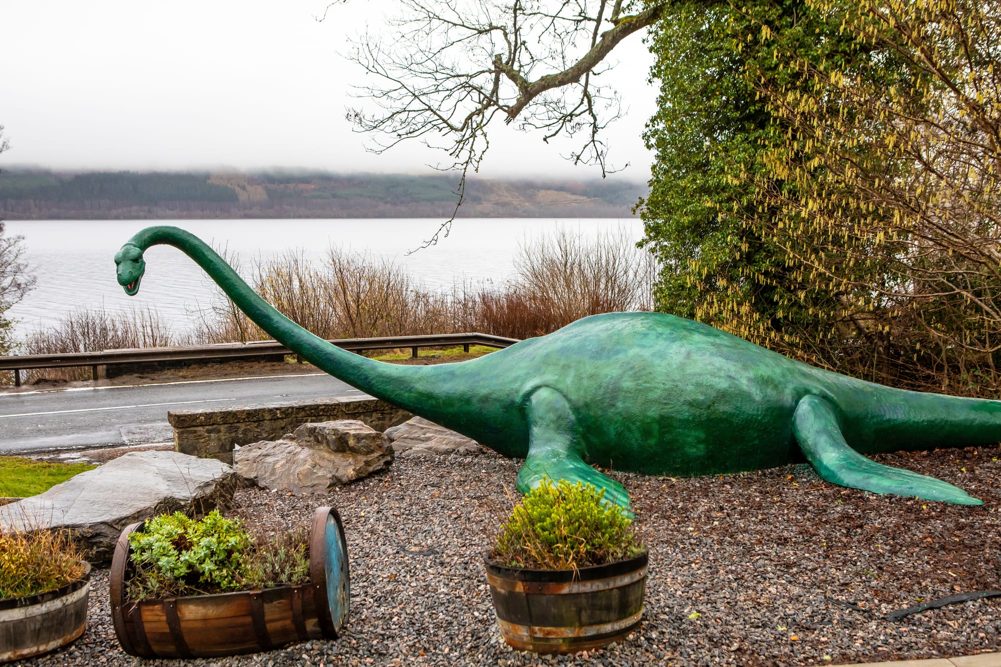 A large, green statue of the Loch Ness Monster on the banks of the loch. The sky is grey and cloudy.