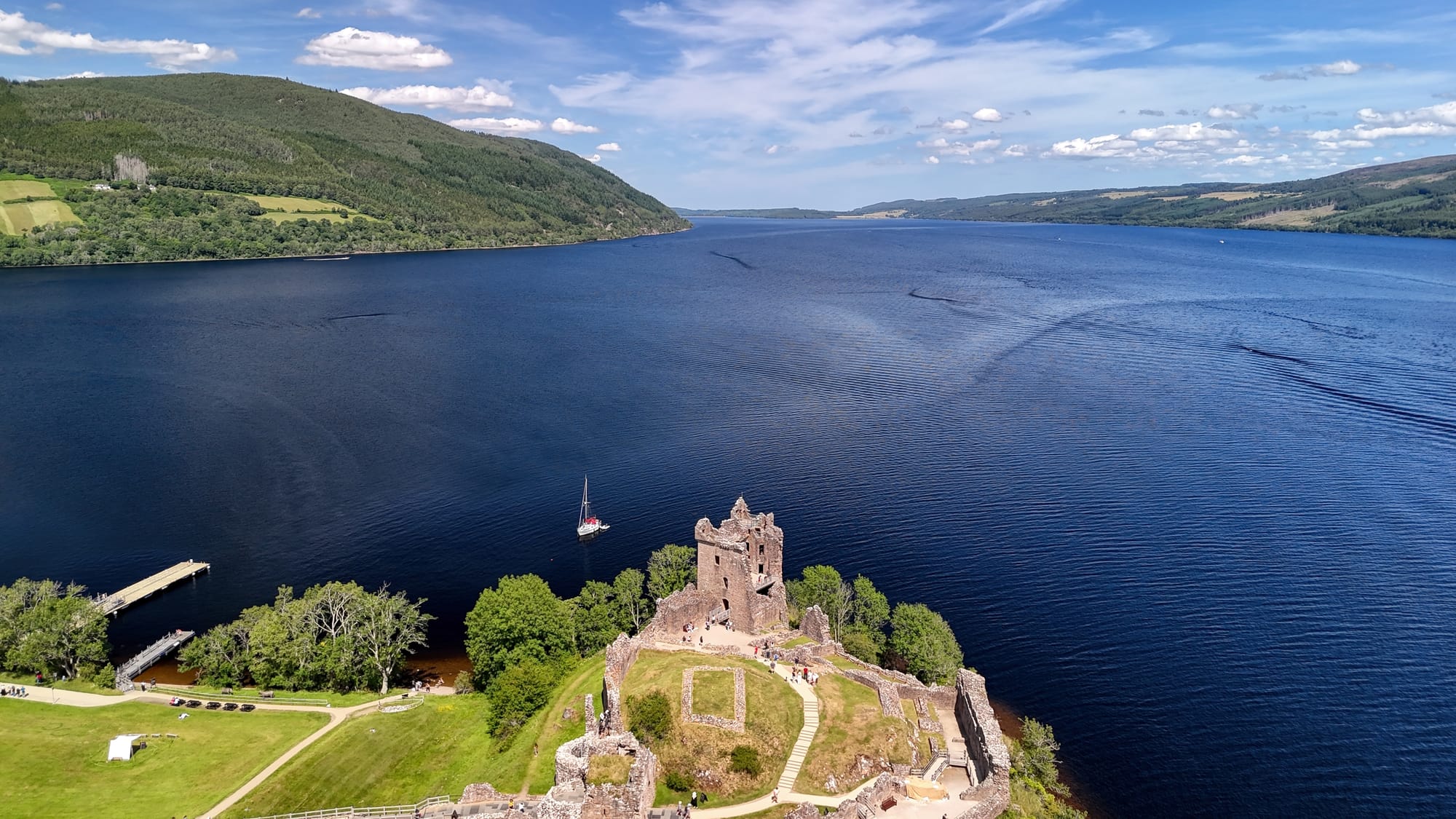 An aerial view of Urquhart Castle on the banks of Loch Ness. The water is a deep blue and the banks are a bright summery green.