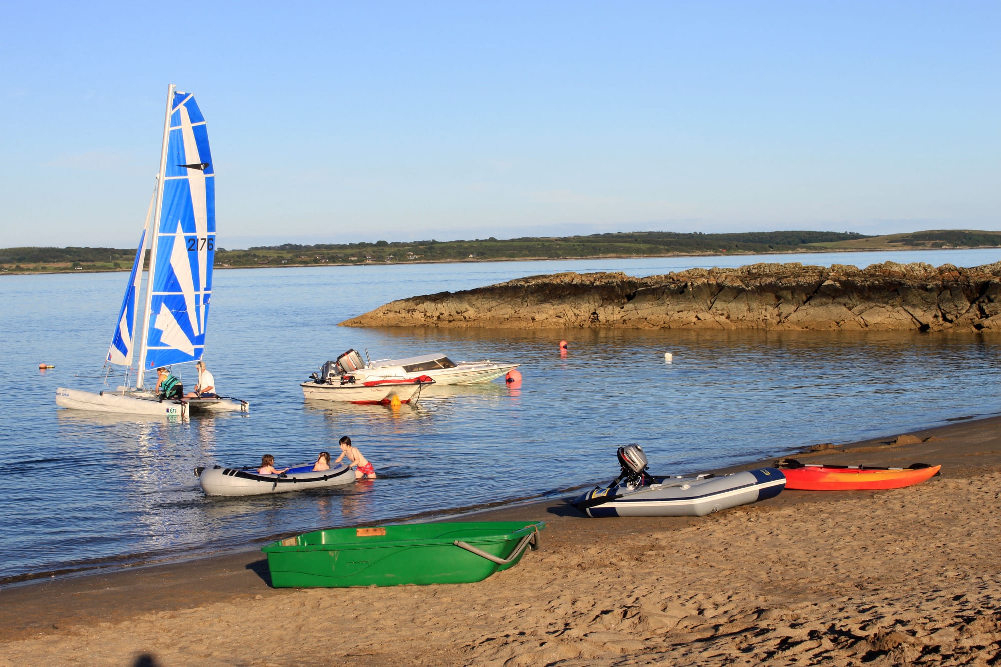 People play on boats near the shore of a sandy beach with a rocky jetty jutting into the water.