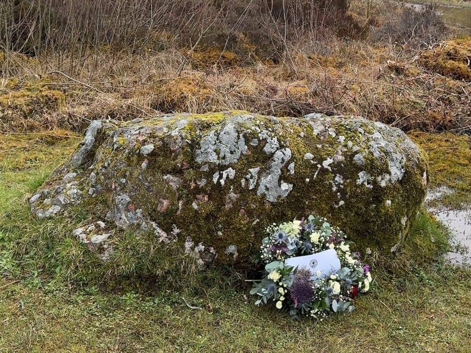 A mossy stone with a floral marker in front of it.