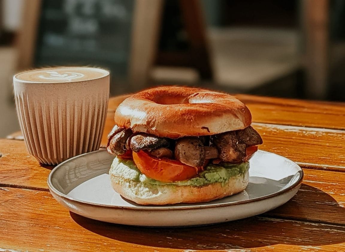 A vegan mushroom bagel with a latte next to it, sat on a wooden table.
