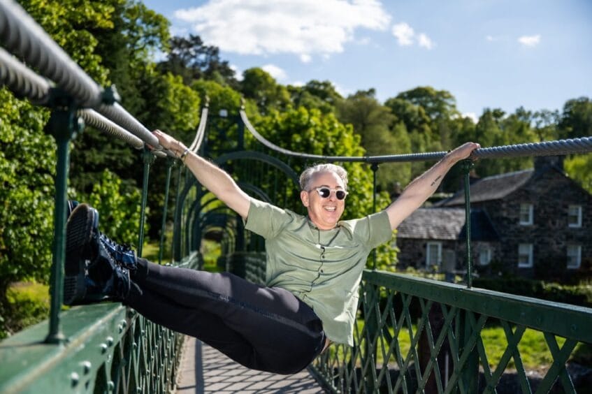Alan Cumming wearing sunglasses with his legs up on the bouncy footbridge.