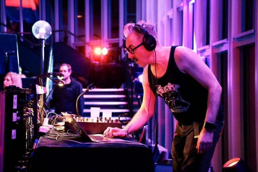 Alan Cumming in a black vest top and headphones, performing a DJ set.