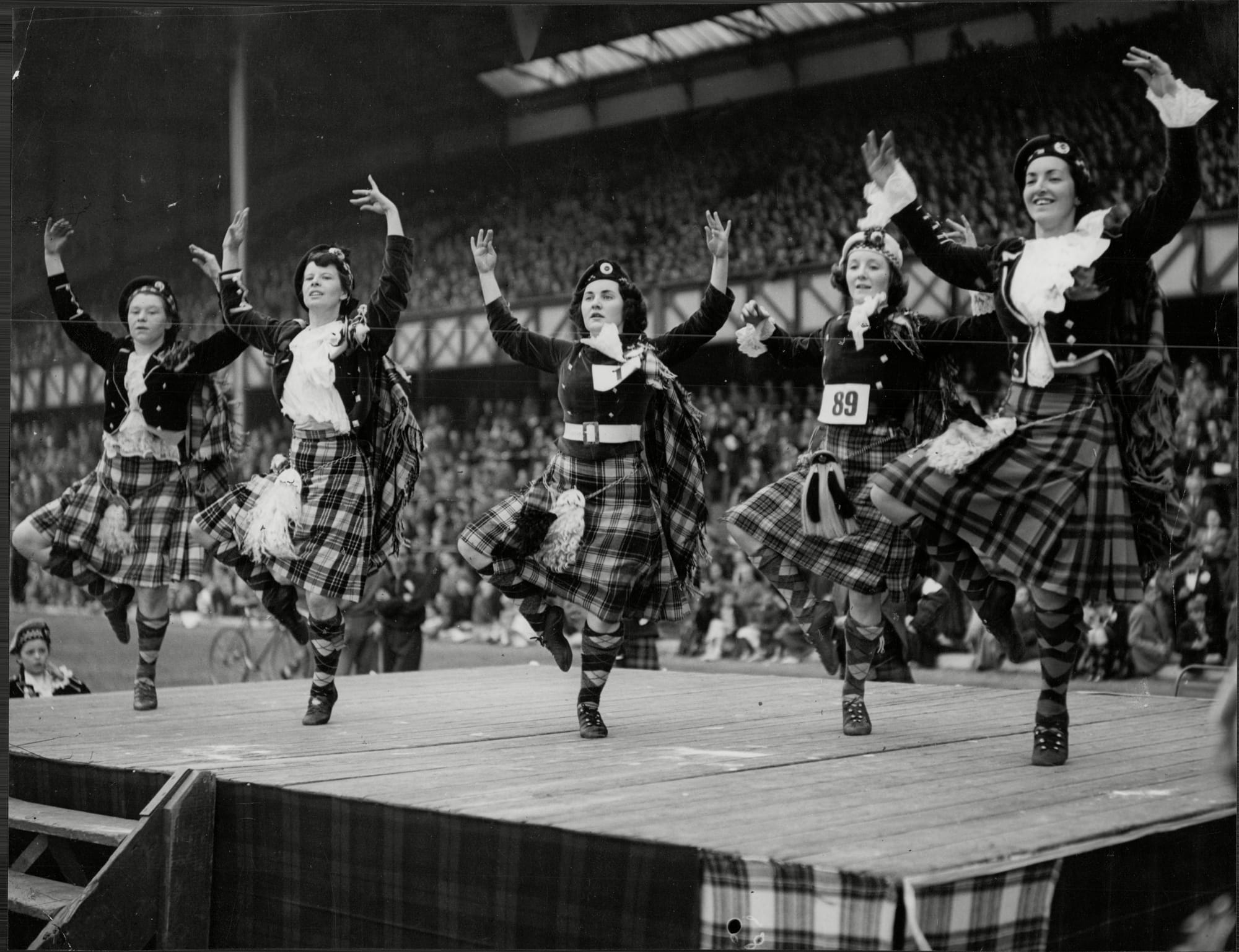 A black-and-white photo of women wearing kilts and Highland dancing on stage.