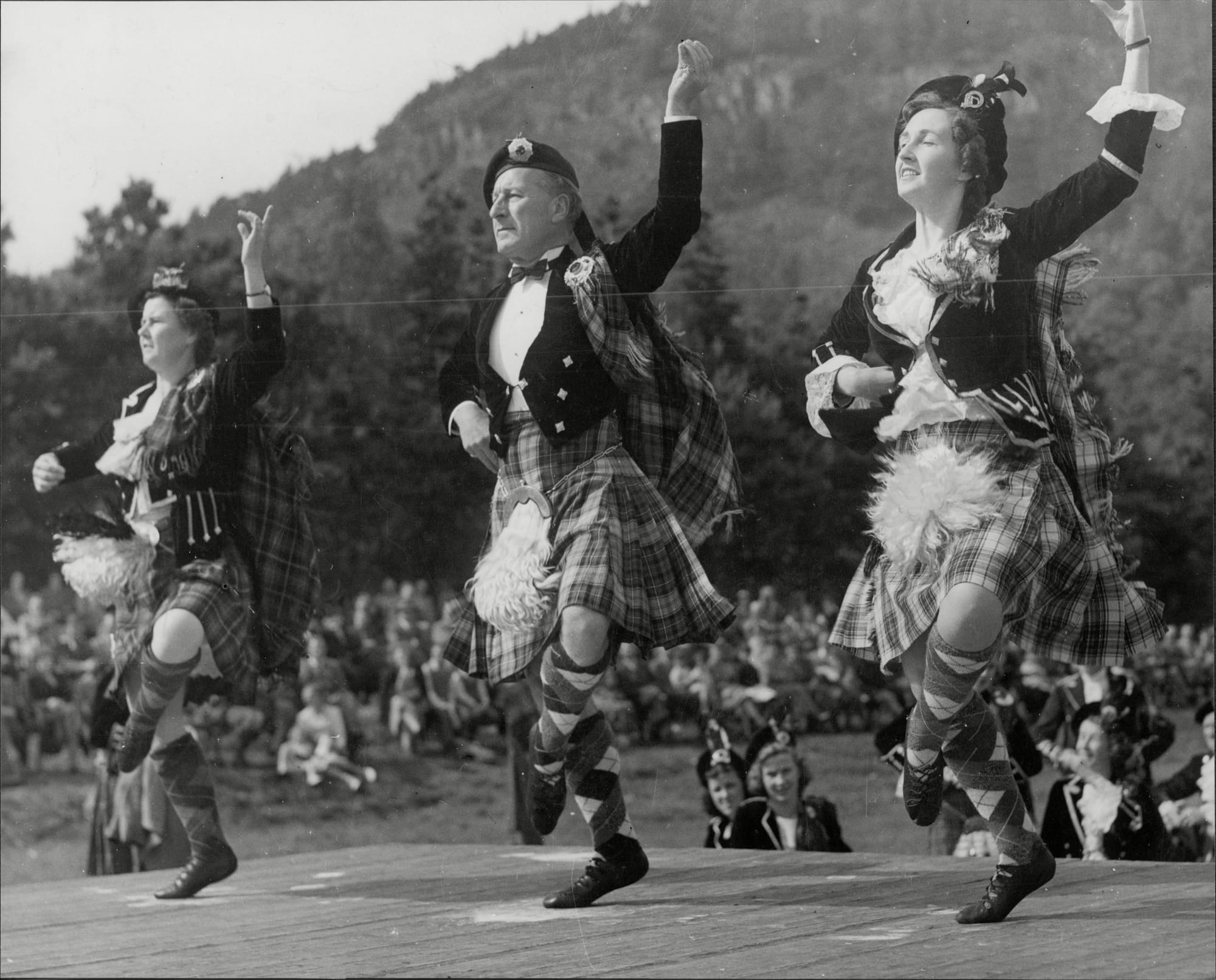 A black-and-white photo of a man and two women competing together on stage in kilts.