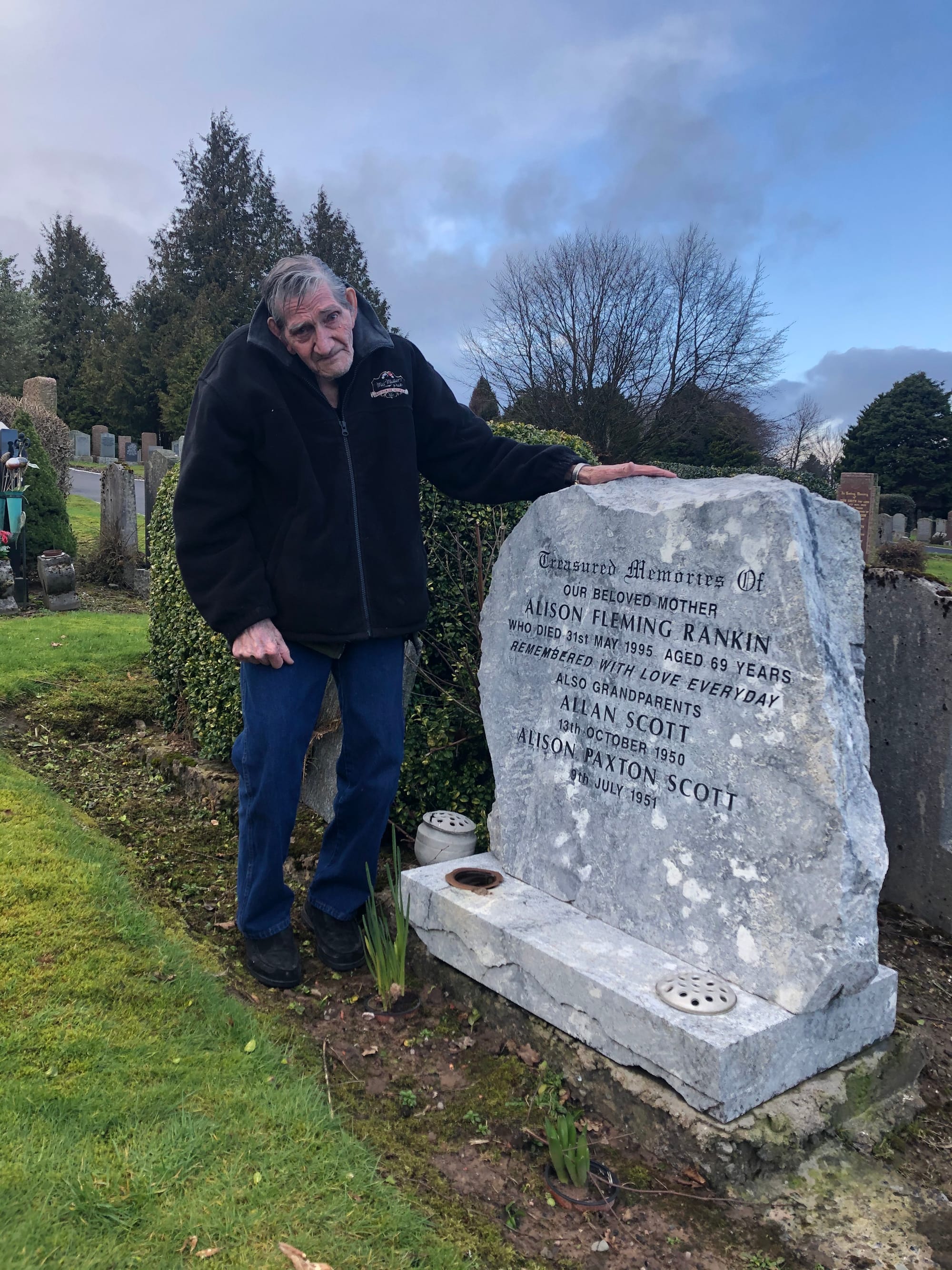 83 year old man stands sadly by a gravestone.